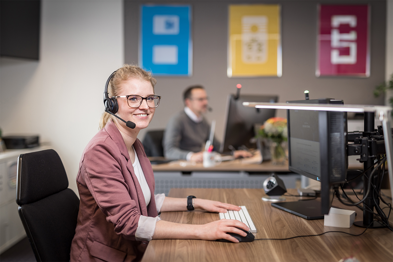 Büroraum; Frau mit Headset im Fokus; im Hintergrund Mann mit Headset